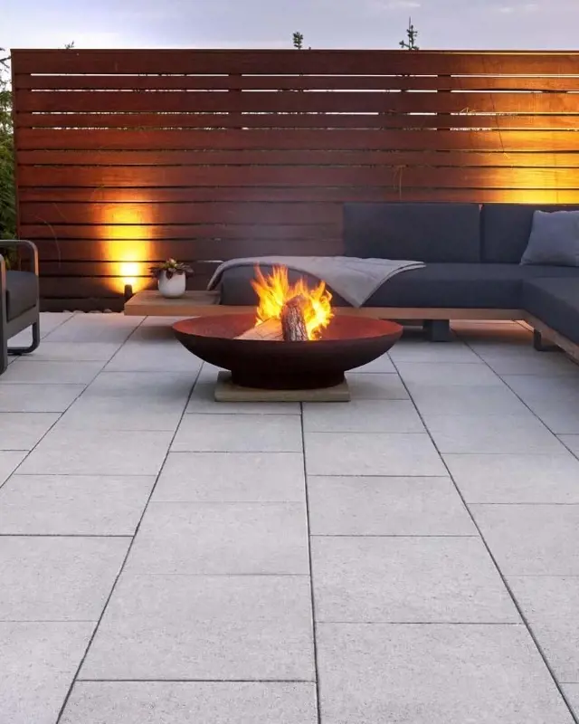 Patio moderne en dalles grises lisses avec foyer central et canapé d’extérieur près d’un écran en bois éclairé. 