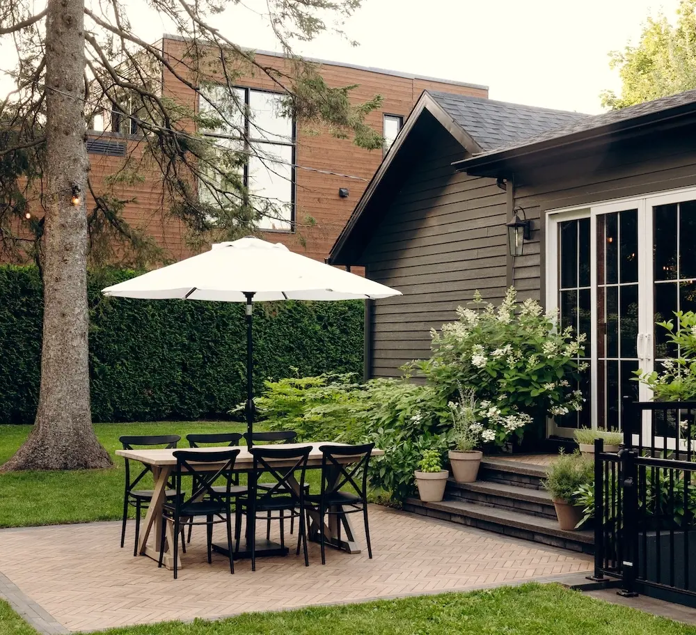 Backyard dining patio with Techo-Bloc Westmount pavers, Limestone cap steps, umbrella, black dining furniture, mature tree, and lush planting.