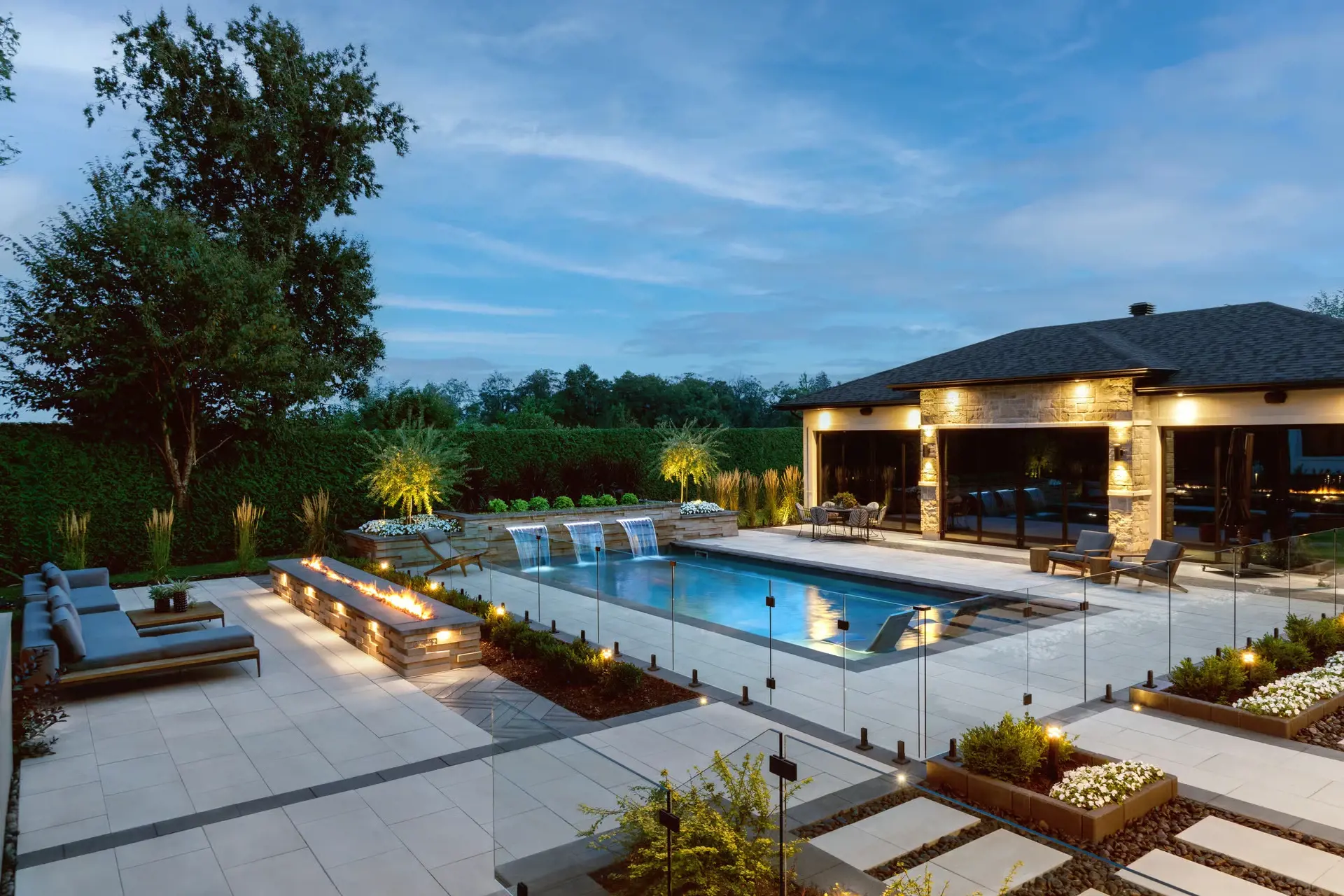 Poolside landscape with expansive light-toned paving, glass fencing, and surrounding greenery.