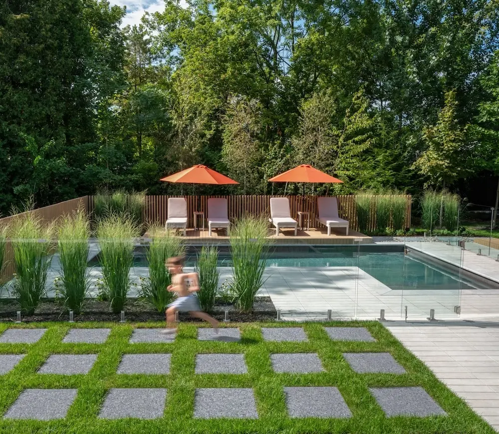 Modern backyard pool area with Techo-Bloc Industria pavers, Terrazzo slabs, grass joints, glass pool fence, lounge chairs, orange umbrellas, and ornamental grasses.