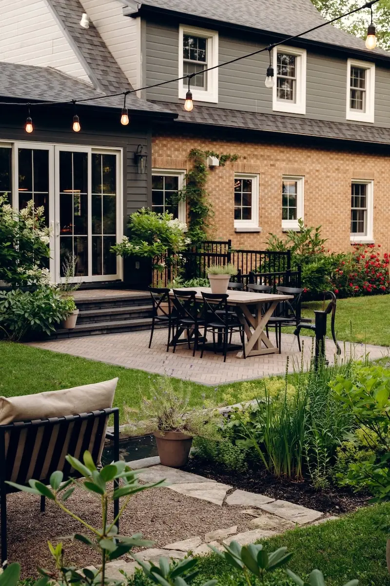 Backyard patio with Techo-Bloc Westmount pavers, Limestone cap edging, outdoor dining furniture, garden beds, string lights, and a brick home exterior.
