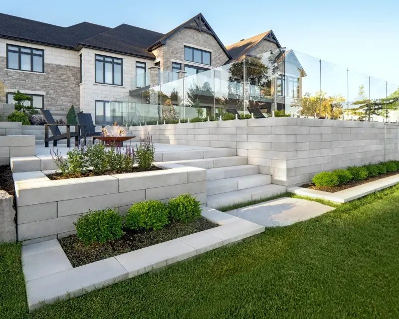 Cour arrière moderne avec des murs de soutènement à plusieurs niveaux en mur Raffinato Lisse de Techo-Bloc, avec marches et garde-corps en verre pour un style architectural raffiné.
