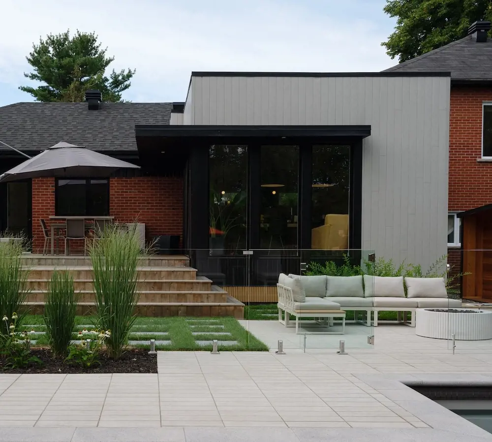 After photo of a modern backyard renovation with Techo-Bloc pavers, patio lounge area, outdoor dining space, garden beds, steps, and a contemporary home extension.
