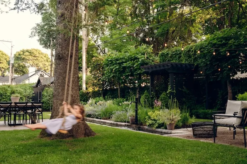Peaceful backyard garden with lawn, mature trees, lounge seating, water feature, and lush greenery.