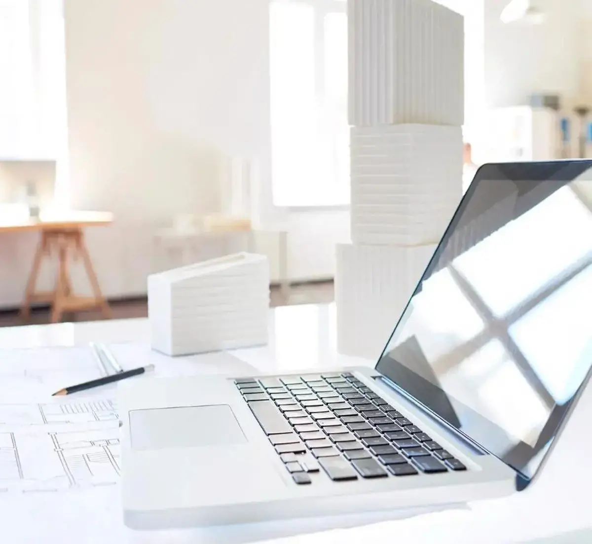 Architectural workspace with a laptop, scale models, and printed plans on a bright desk.