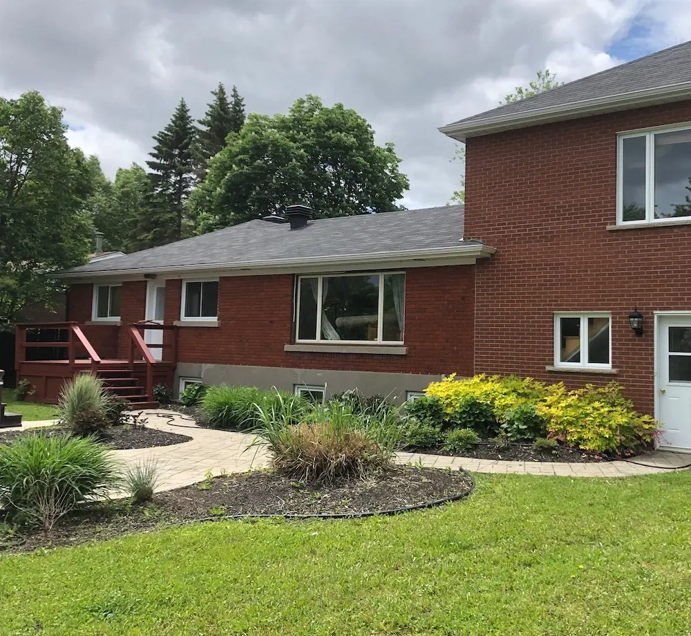 Before photo of a brick home backyard with a curved garden bed, lawn, shrubs, and an older patio area before renovation.