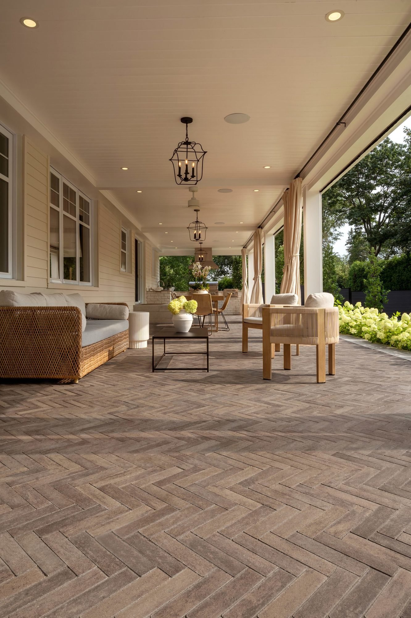 Espace de vie extérieur avec pavés Westmount couleur Brun Châtaigne sous un patio couvert comprenant mobilier tressé et rideaux légers.