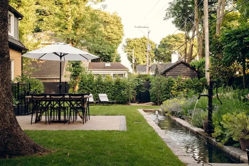 Renovated family backyard with Techo-Bloc Westmount pavers, Limestone cap edging, a lawn, water feature, outdoor dining area, umbrella, and lush garden planting.