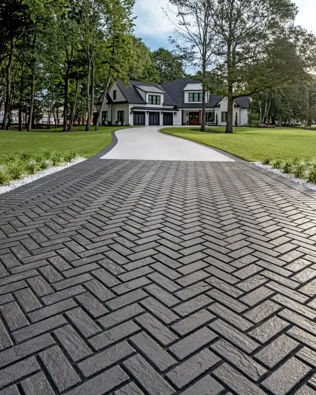 Elegant driveway paved with Techo‑Bloc Mika Paver in onyx black, arranged in a herringbone pattern surrounded by lush greenery.