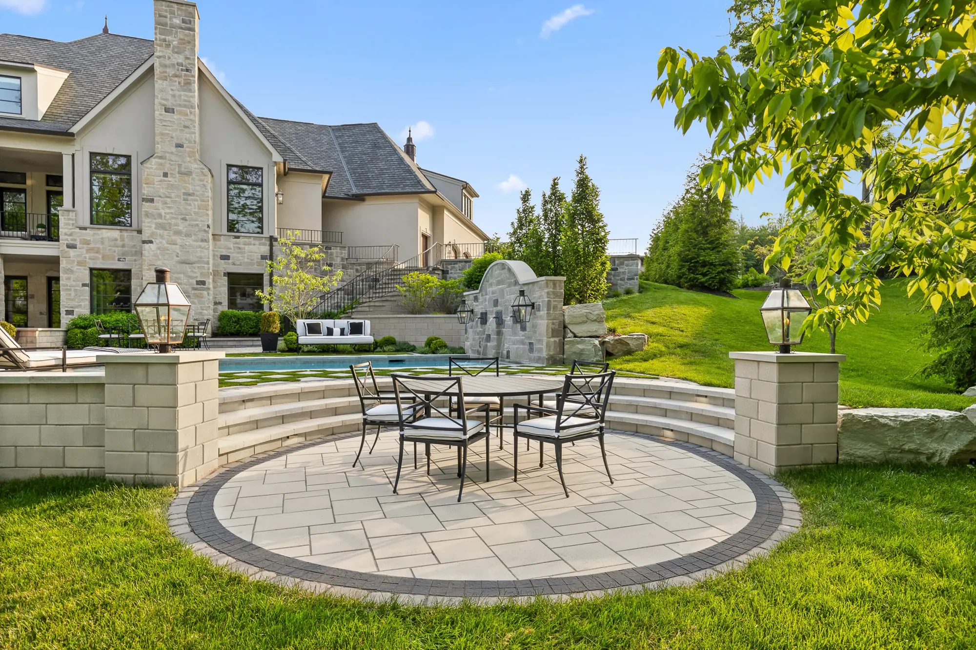 Maison de luxe en pierre et stuc avec terrasse circulaire en pavés, table ronde, piscine et mur-fontaine dans un jardin paysager.