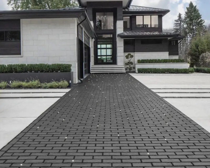 Maison moderne avec une longue allée en pavés Mika de Techo-Bloc disposés en motif modulaire sombre, bordée de haies basses menant à l’entrée.