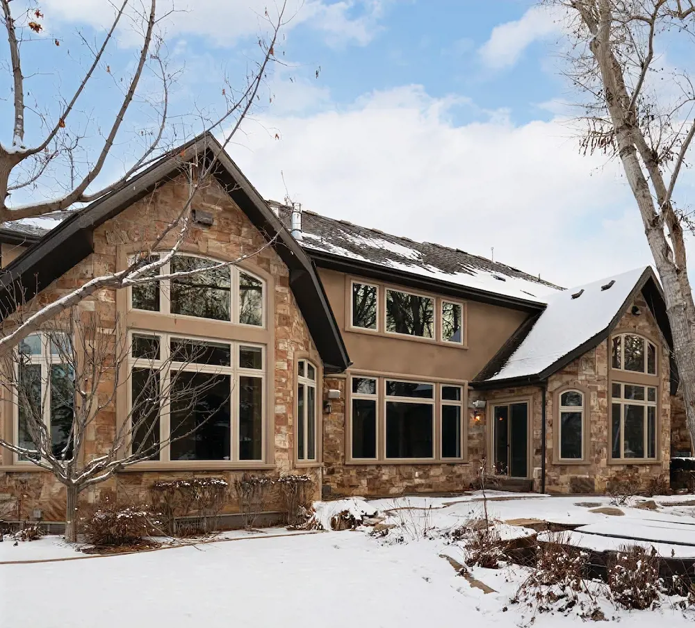 Before photo of a traditional stone home exterior in winter with large windows, bare trees, snow, and an unfinished front landscape.