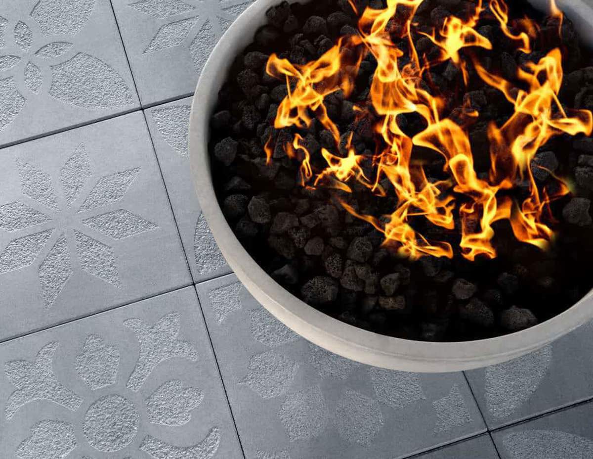 A bowl of gray rock with a calm fire within on pavers also gray where flowers of different style can be seen. 