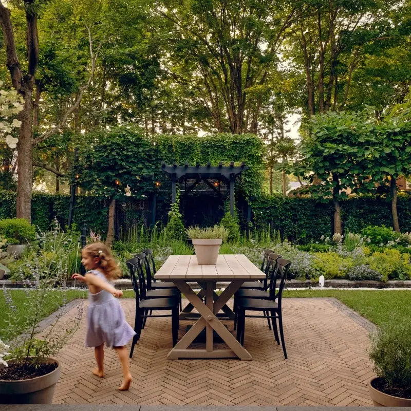 Outdoor dining area with Techo-Bloc Westmount pavers in Chestnut Brown laid in a herringbone pattern, surrounded by garden greenery.