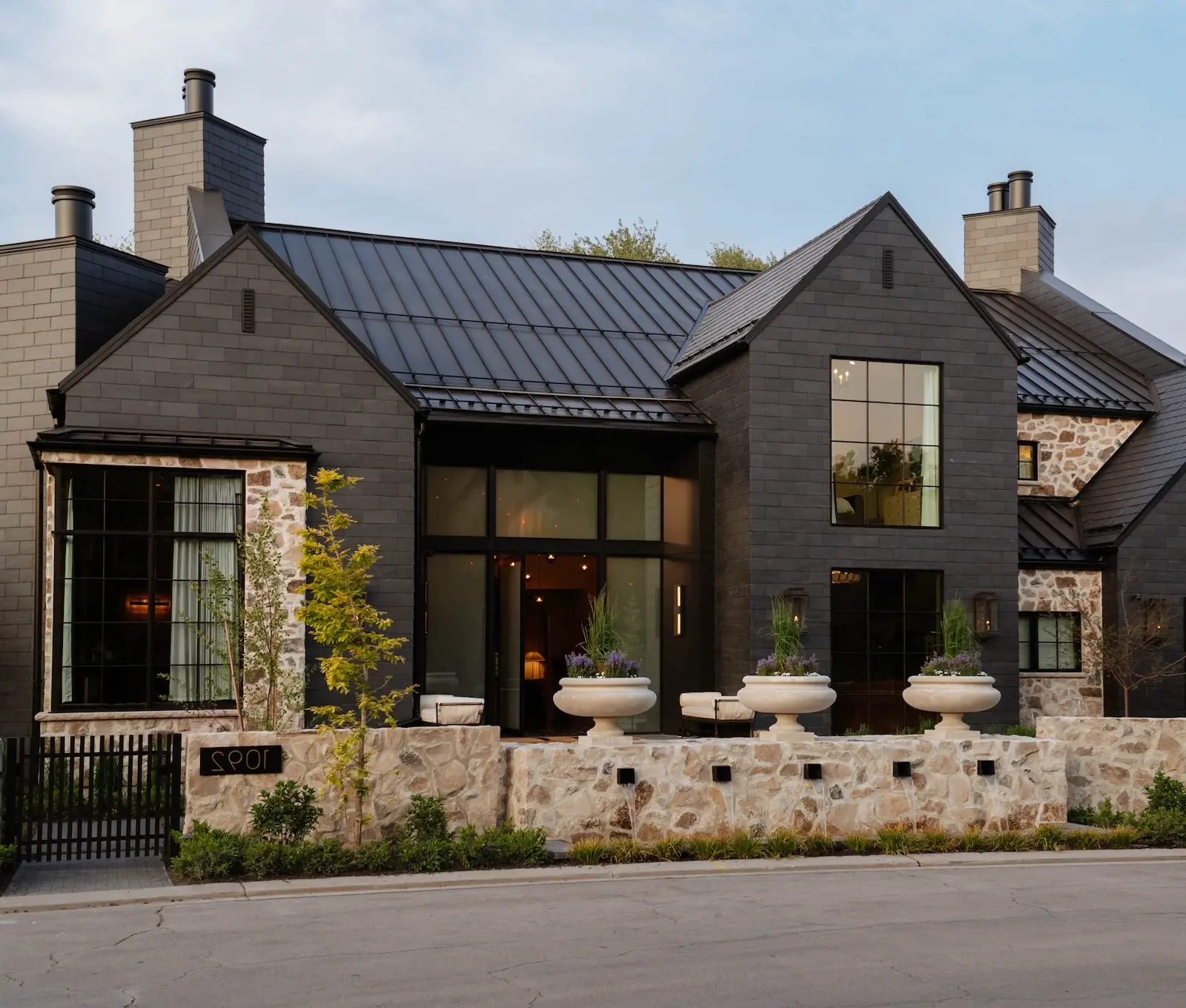 After photo of a renovated luxury home exterior with modern dark siding, stone façade, black metal roof, front wall planters, and refined curb appeal.