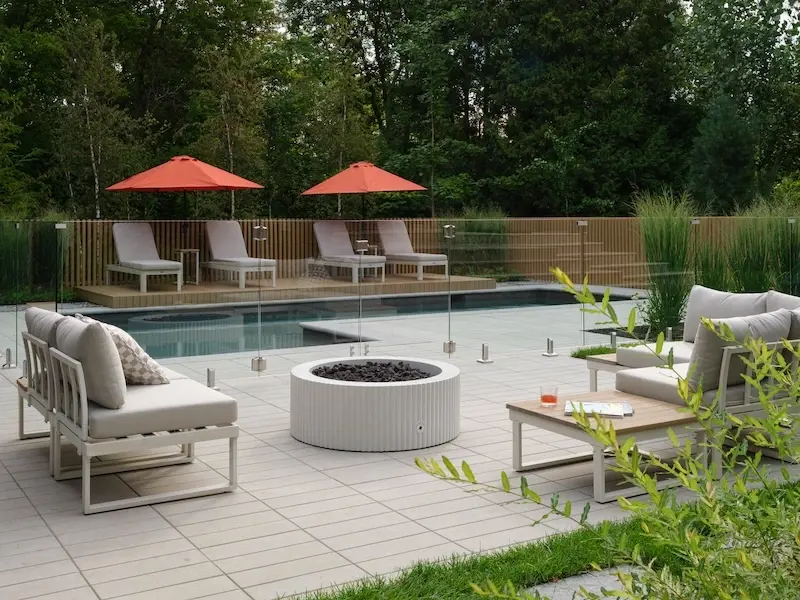 Backyard pool lounge area with Techo-Bloc Industria pavers, outdoor sofas, round fire feature, lounge chairs, umbrellas, and glass pool fence.