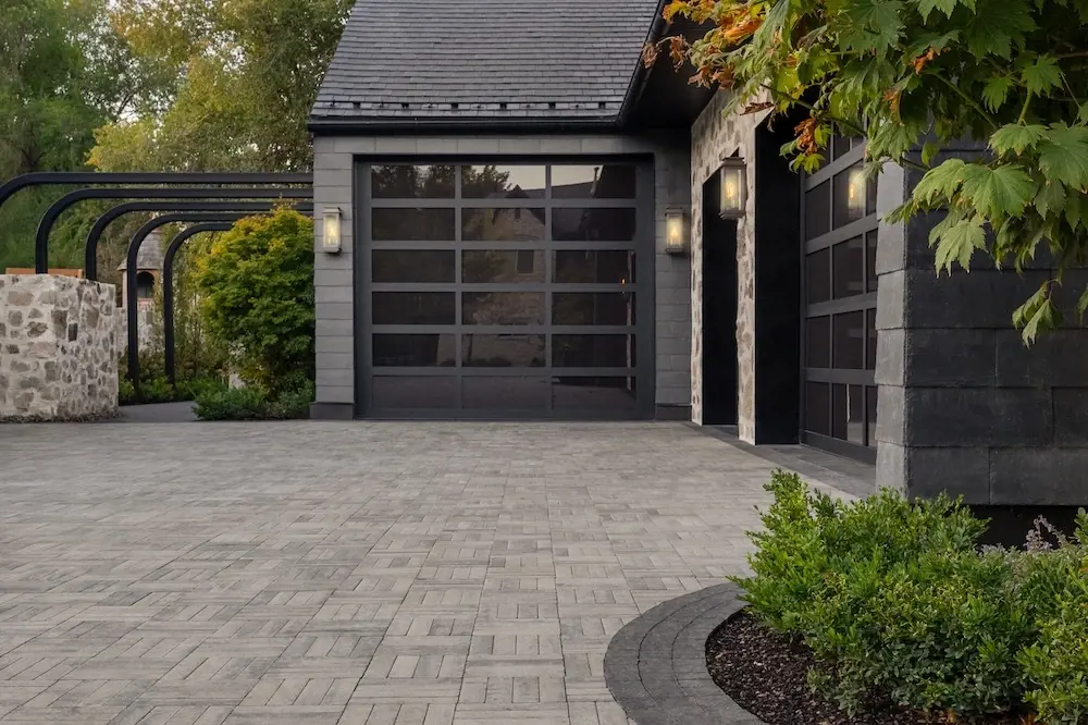 Modern driveway with Techo-Bloc Westmount pavers, dark garage doors, stone wall details, curved edging, and foundation planting.