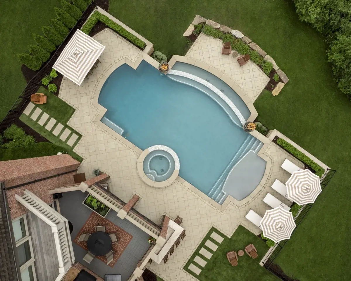 Vue aérienne d’une cour arrière haut de gamme avec piscine creusée sur mesure, spa intégré et grande terrasse en pavés de pierre entourée d’aménagement paysager.