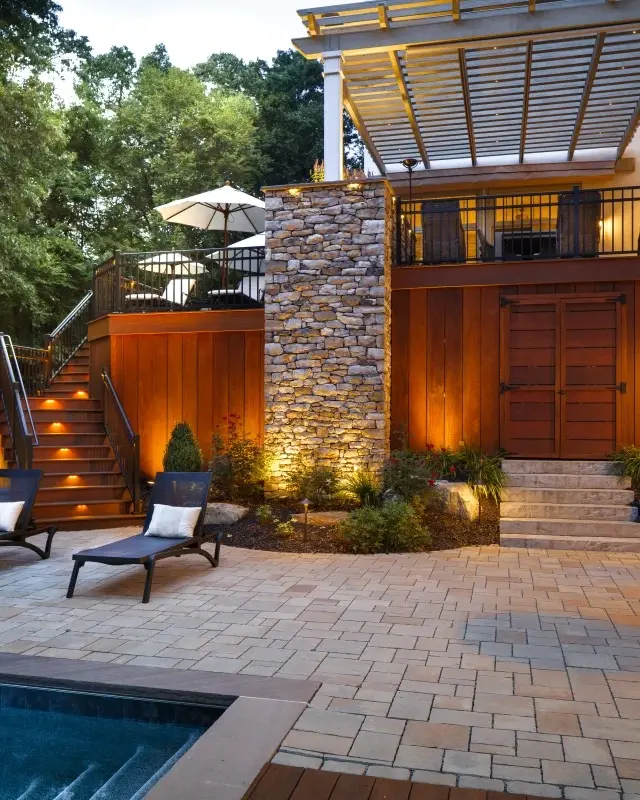 Poolside patio project with beige modular slabs by Techo‑Bloc, featuring a pergola, stone chimney, and soft evening lighting. 