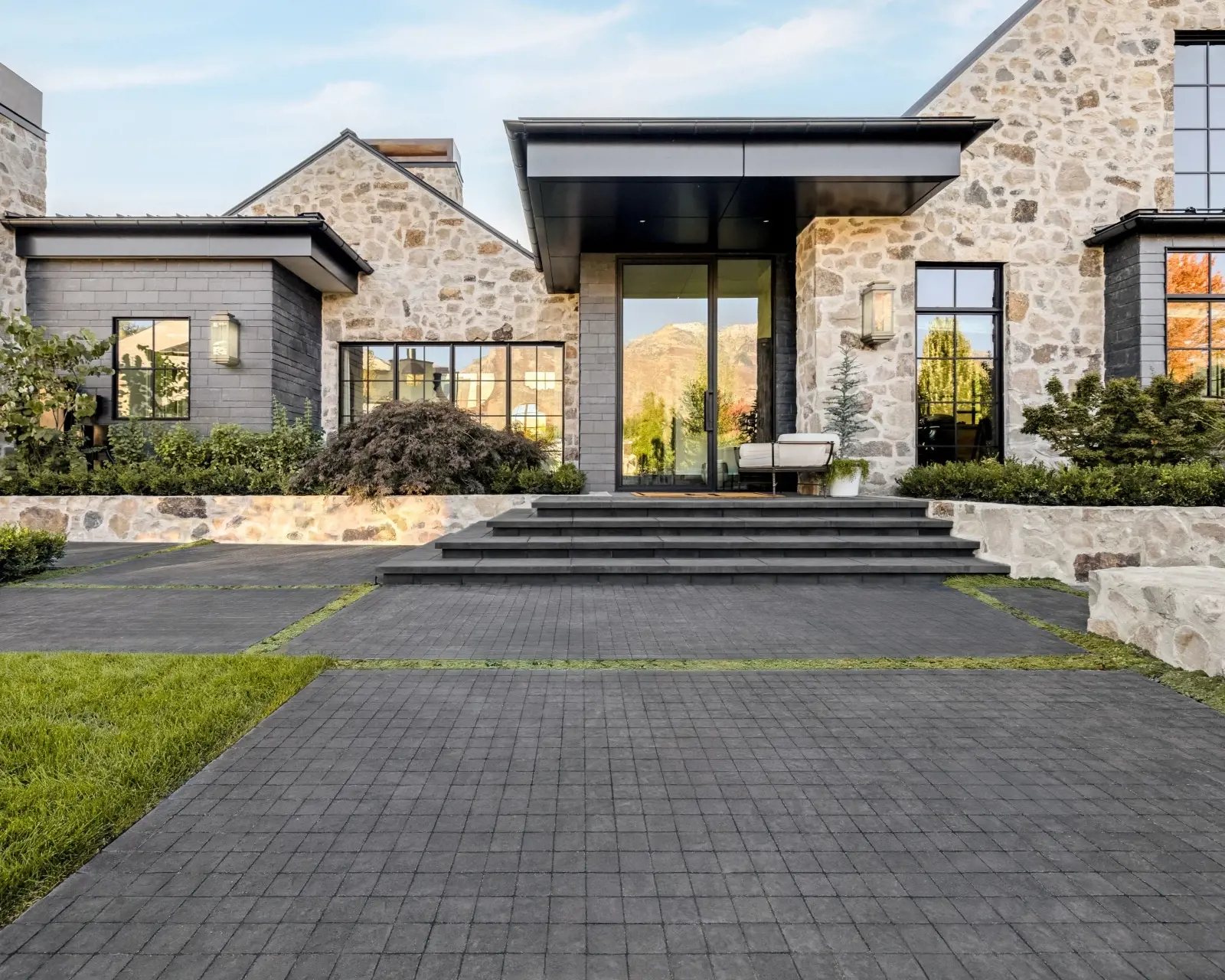 Contemporary home exterior featuring textured stone walls, layered landscaping, and a dark-toned driveway that adds depth, warmth, and a sense of permanence.