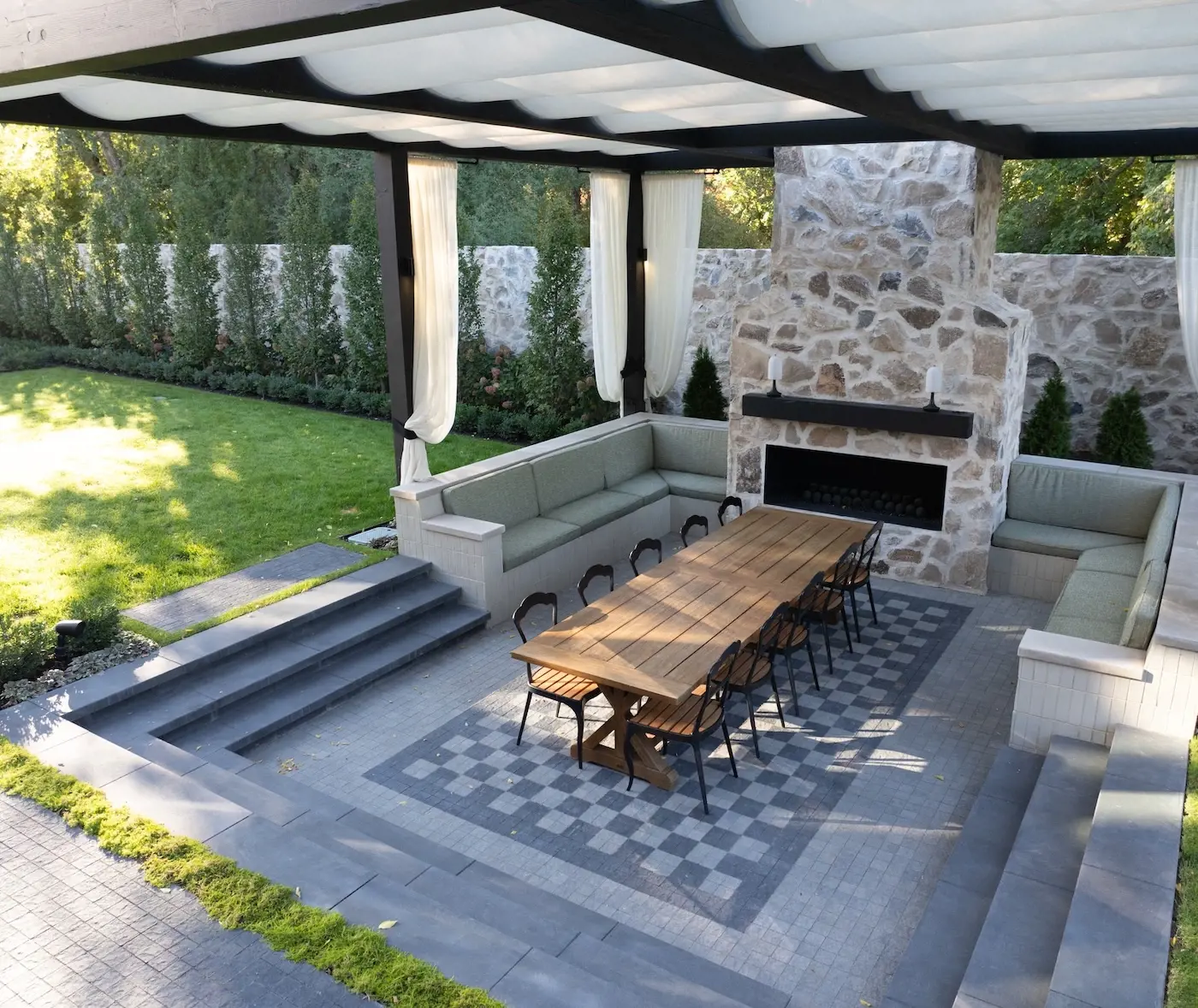 Covered outdoor dining area with Techo-Bloc Squadra pavers, stone fireplace, built-in seating, large wood table, and garden views.