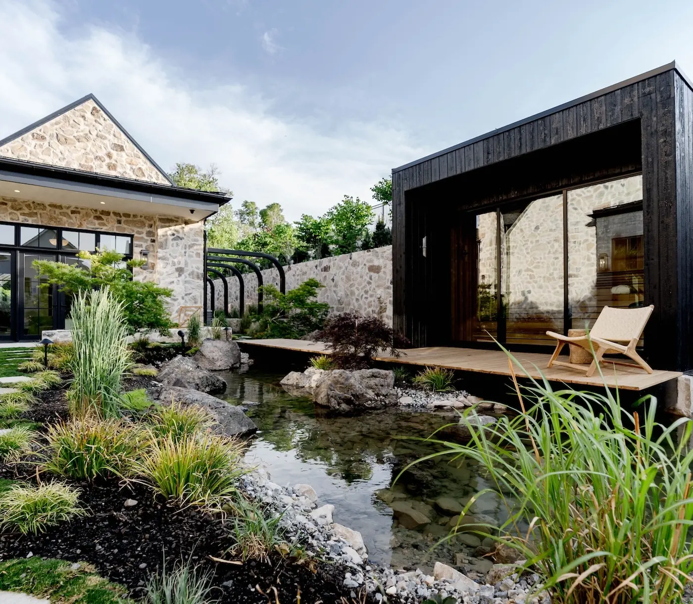 Luxury backyard garden with natural pond, stone architecture, black modern pavilion, ornamental grasses, boulders, and layered planting.