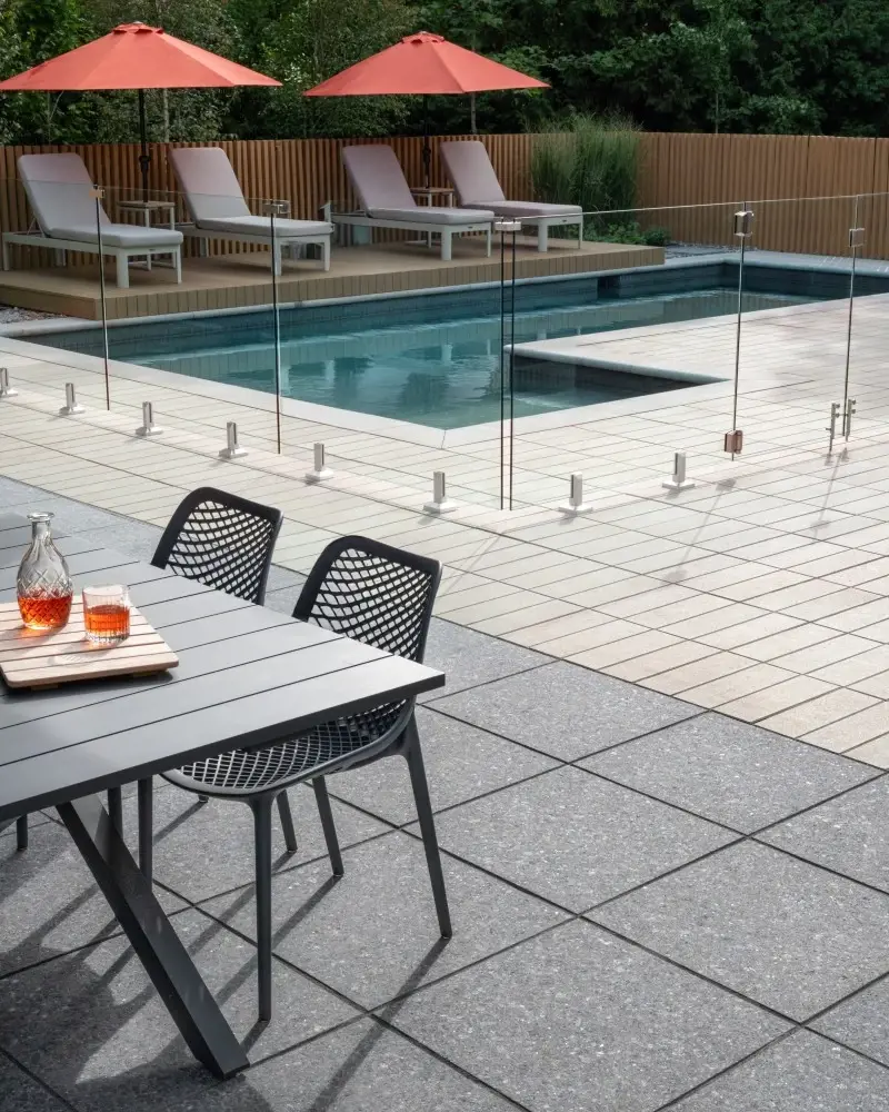 Modern backyard pool with light concrete pavers, dark accent slabs, and glass railing creating a controlled contrast design.