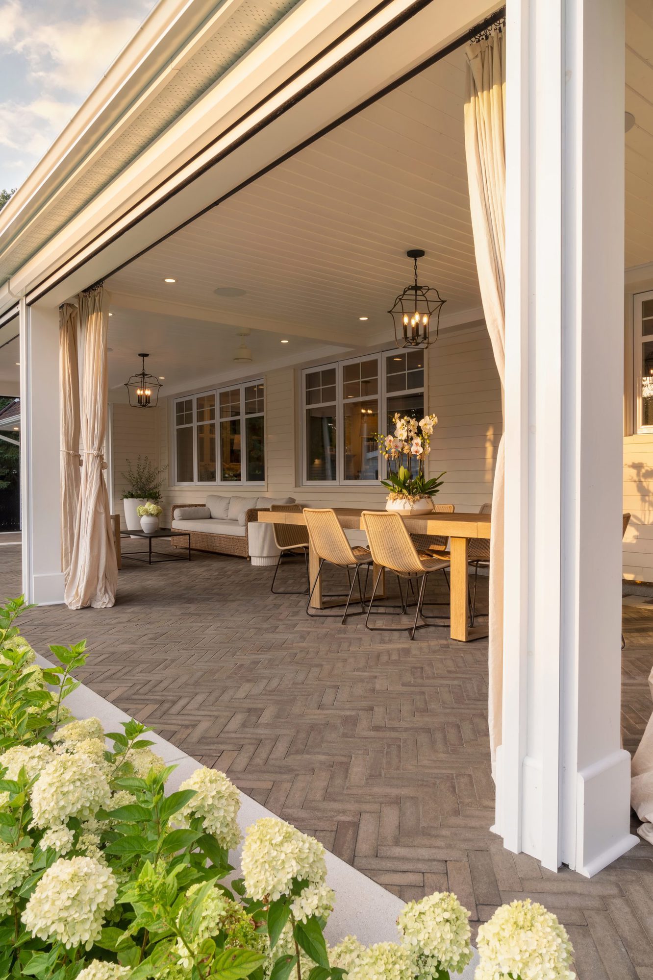 Espace repas extérieur couvert avec rideaux légers fluides, mobilier tressé et textures boisées sur des pavés Westmount.