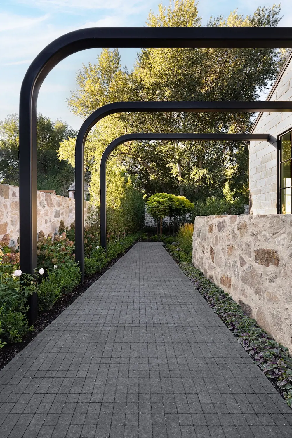 Elegant garden walkway with Techo-Bloc Squadra pavers, black metal arches, stone walls, and layered greenery.