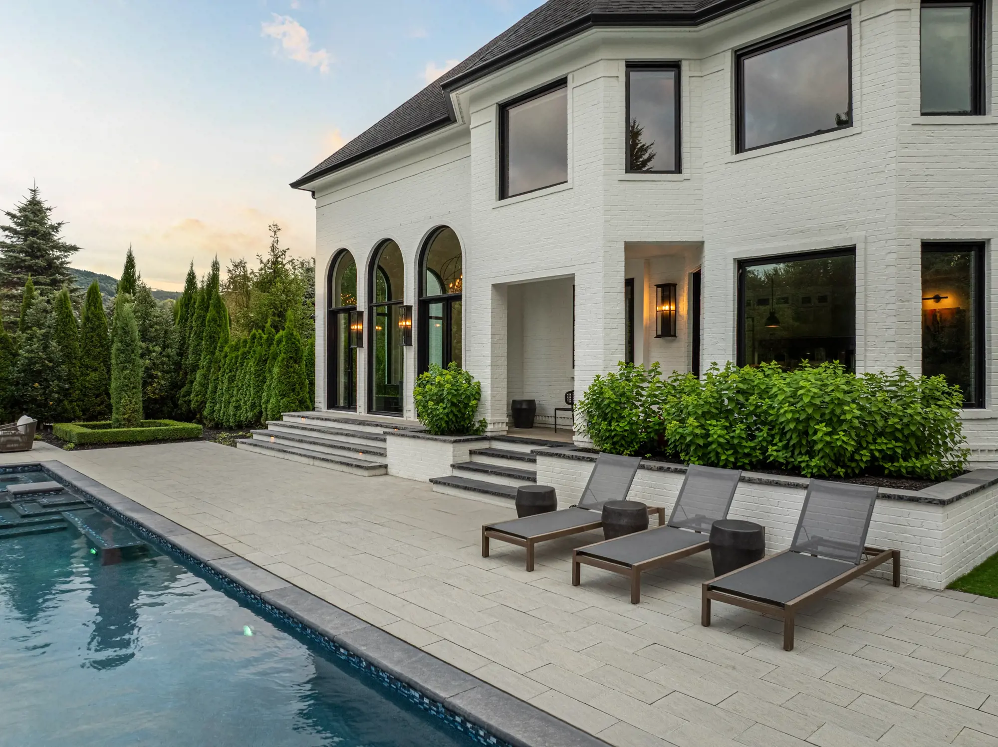 Maison de luxe en brique blanche au crépuscule, aux hautes fenêtres cintrées noires, avec piscine et chaises longues grises sur une terrasse en pavés.