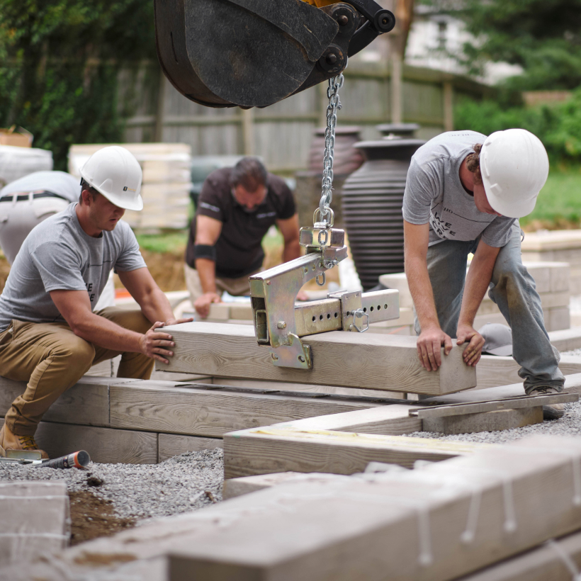 Équipe d’aménagement paysager installant des blocs de béton à l’aide d’un équipement de levage sur le chantier d’un projet résidentiel d’aménagement extérieur, utilisant des produits Techo-Bloc.