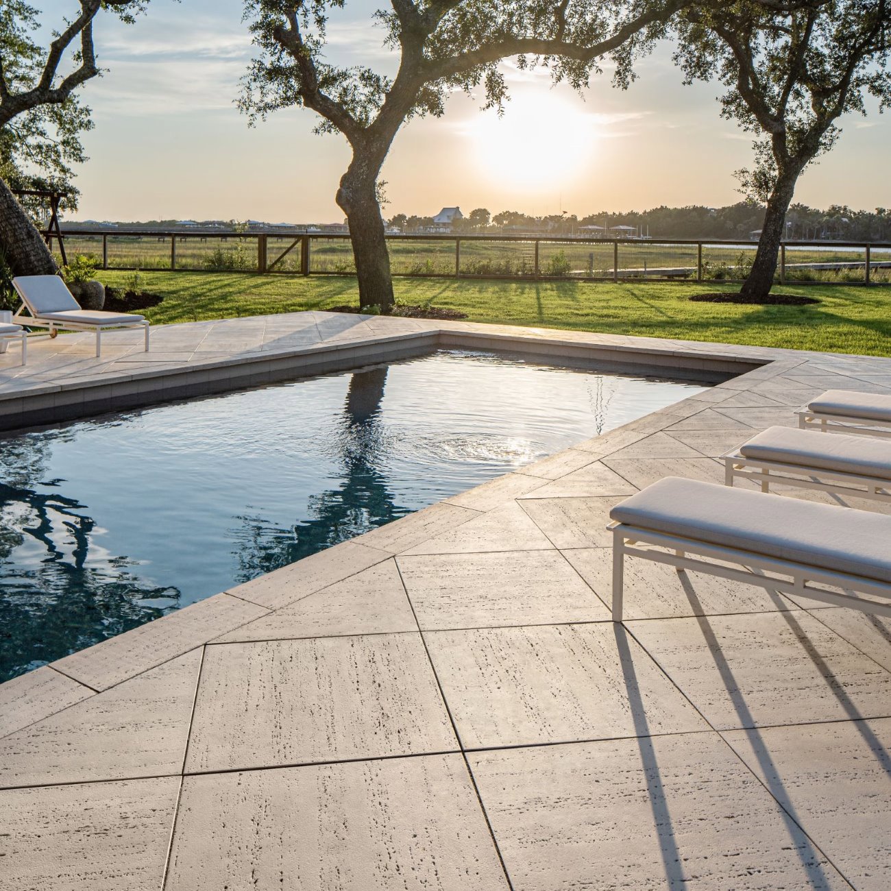 Travertina Raw Slabs by Techo-Bloc in beige, textured patio surrounds pool with white loungers, open fields, and sunset sky.