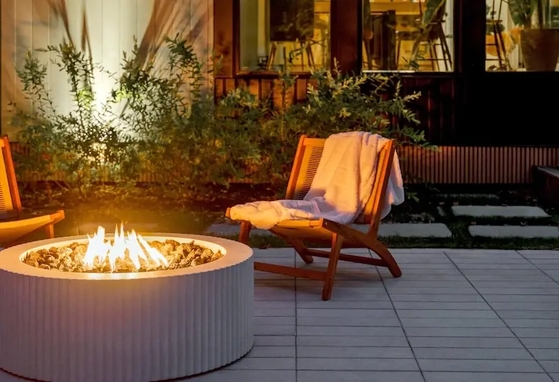 Evening backyard lounge area with Techo-Bloc Industria pavers, Terrazzo slab accents, round fire feature, wood lounge chair, and lush planting.