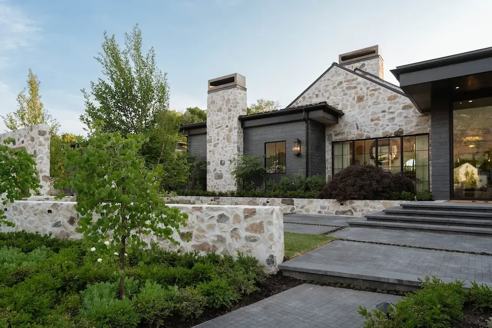 Luxury stone home exterior with Techo-Bloc Squadra pavers, layered garden planting, low stone walls, and wide front steps.