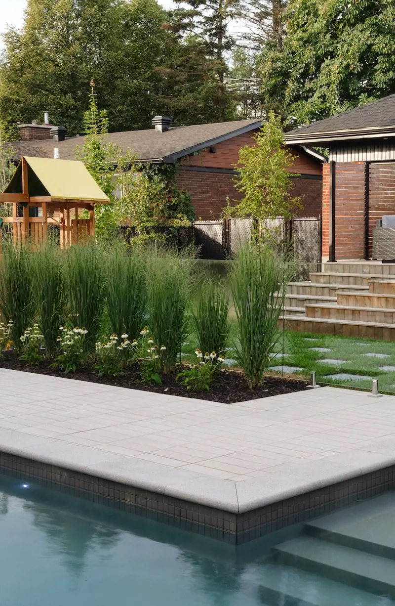 Modern poolside landscape with Techo-Bloc Industria pavers, Terrazzo slab accents, planting beds, ornamental grasses, wood privacy fence, and steps.