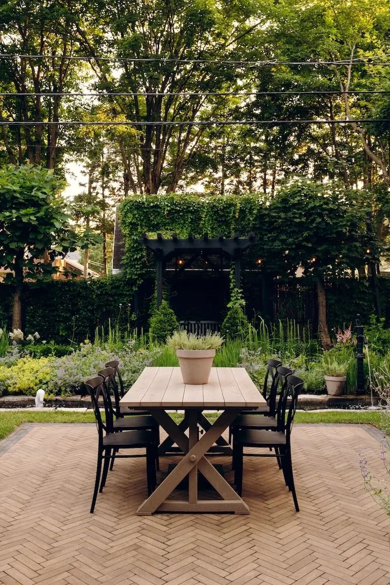 Intimate backyard dining nook with Techo-Bloc Westmount pavers, Limestone cap edging, wood table, black chairs, garden arch, and lush greenery.
