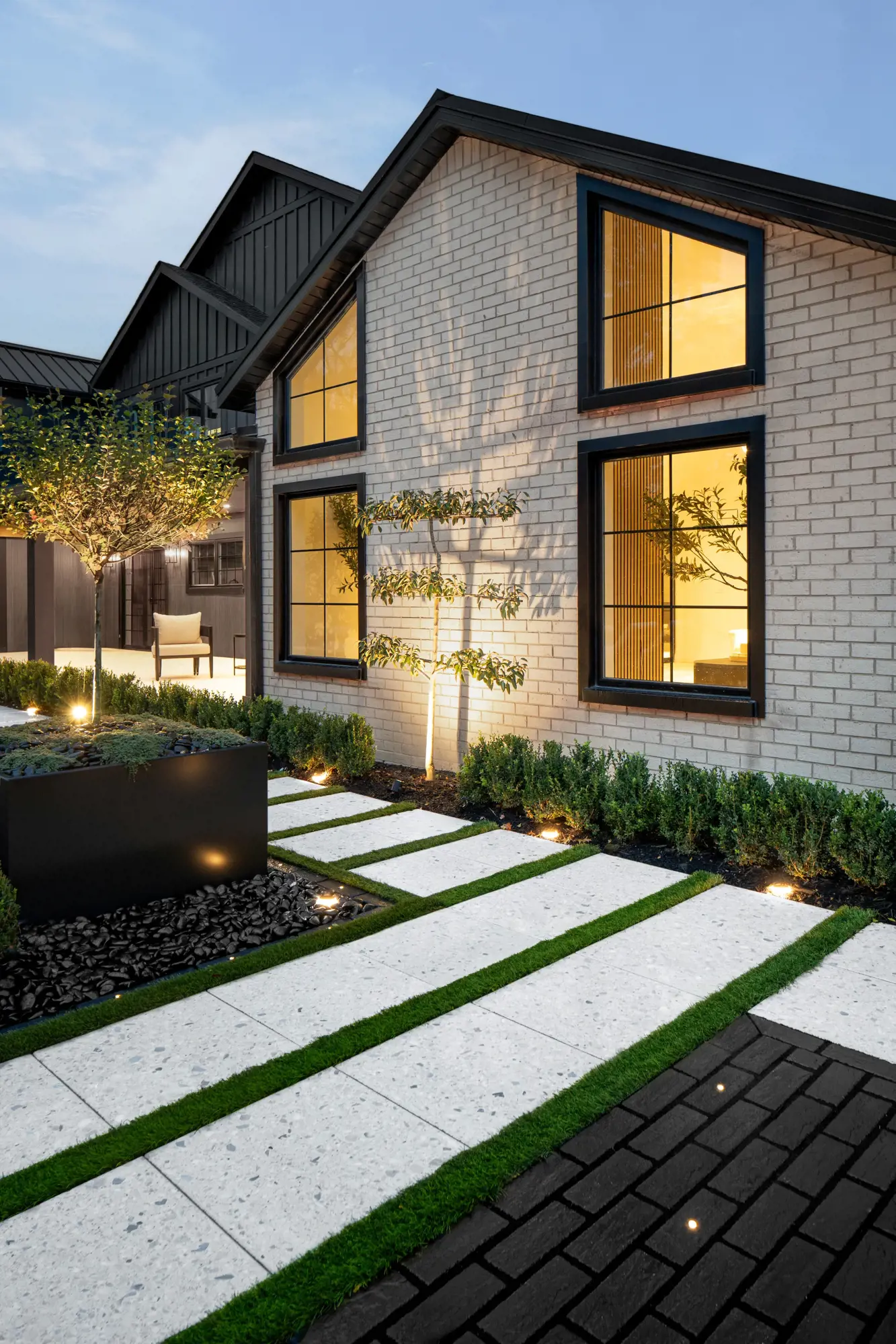 Façade de la maison au crépuscule avec allée en Terrazzo, dalles à joints gazonnés et éclairage paysager chaleureux