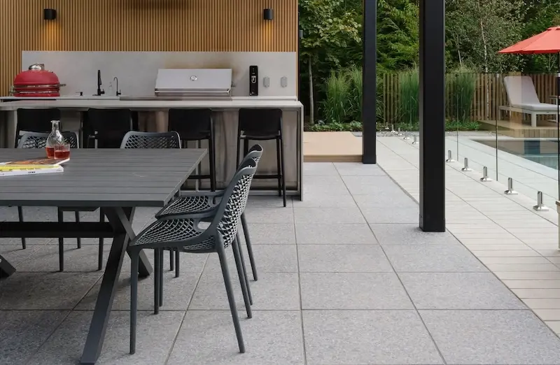 Covered outdoor kitchen and dining area with Techo-Bloc Terrazzo slabs, glass pool fence, black dining furniture, and modern poolside design.