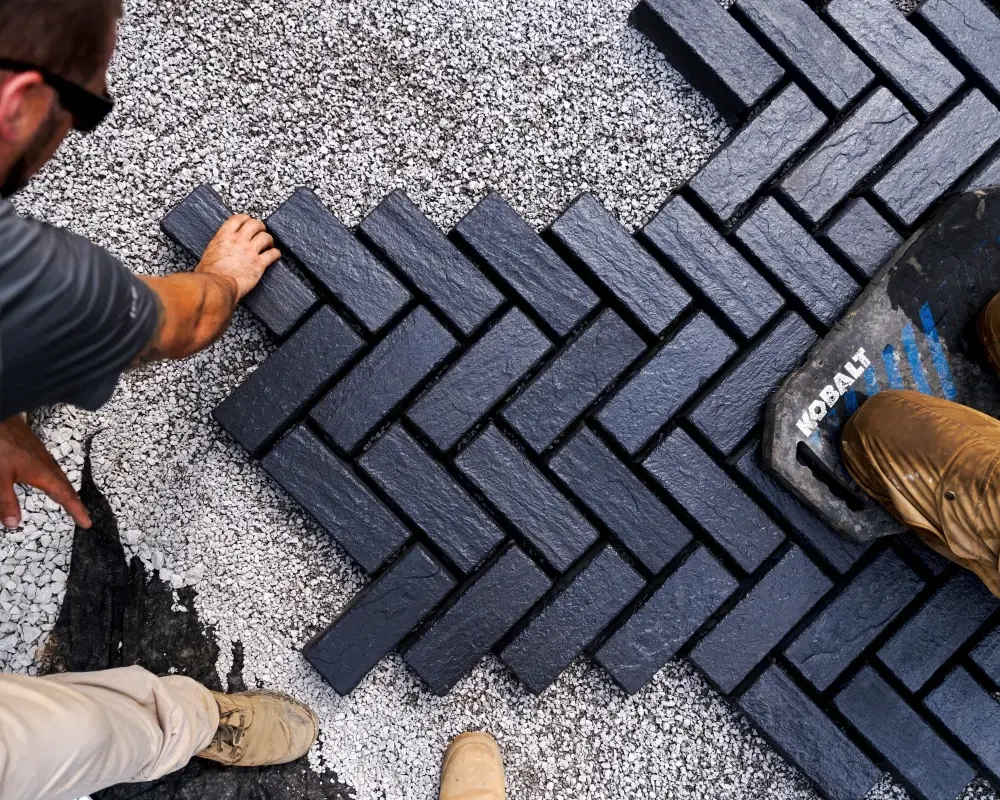 Entrepreneur posant des pavés foncés en motif à chevrons sur une base de gravier, illustrant un travail d’installation précis.