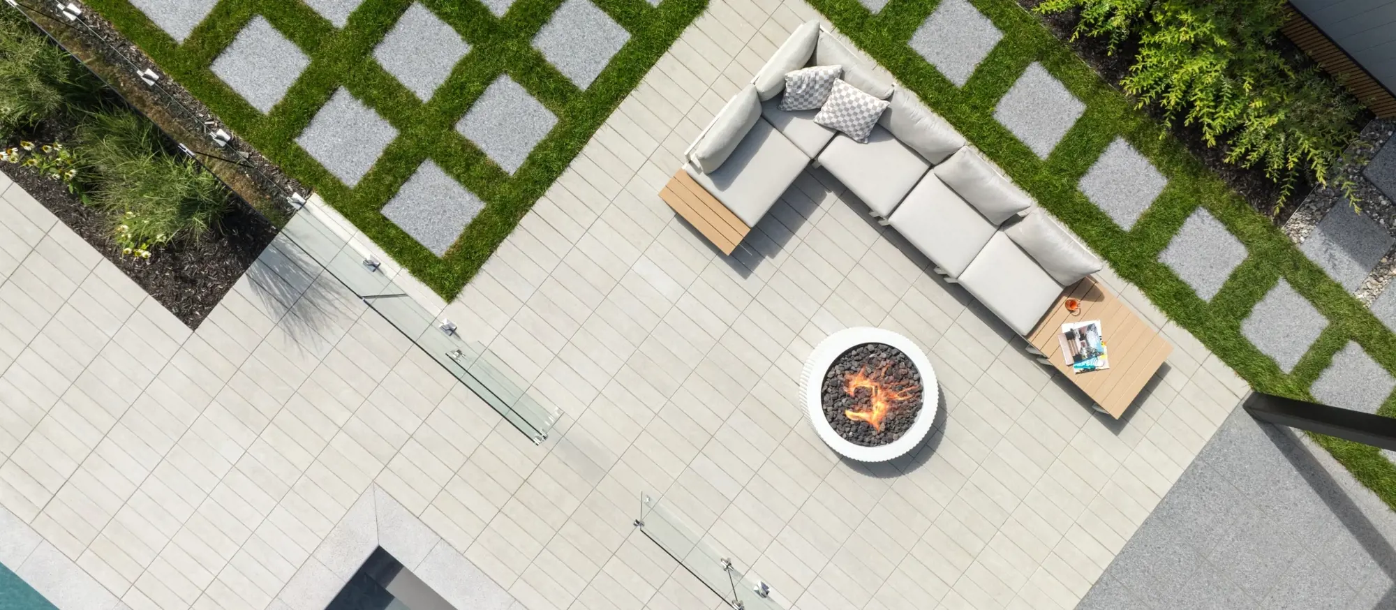 Aerial view of a modern outdoor living space featuring light-toned paving, a circular fire pit, modular seating, and geometric grass inlays.