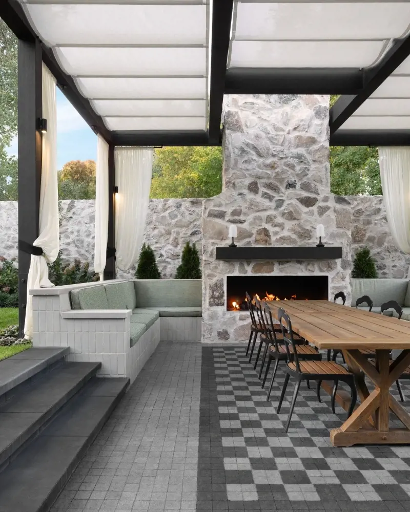 Modern covered patio with grey Techo-Bloc pavers, outdoor dining table, built-in seating, and stone fireplace.
