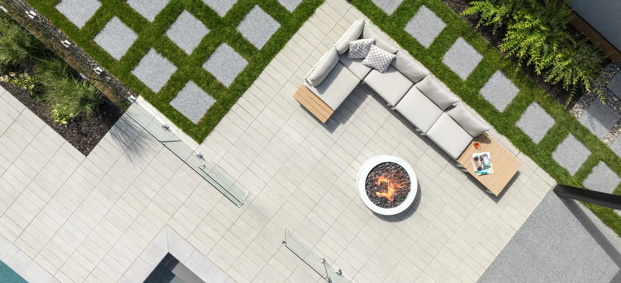 Aerial view of a modern outdoor living space featuring light-toned paving, a circular fire pit, modular seating, and geometric grass inlays.