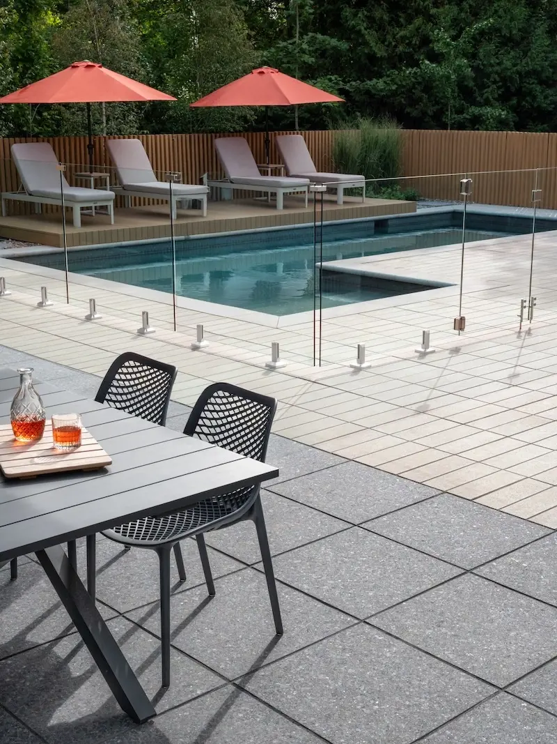 Outdoor dining area beside the pool with Techo-Bloc Industria pavers, Terrazzo slabs, glass pool fence, lounge chairs, and orange umbrellas.