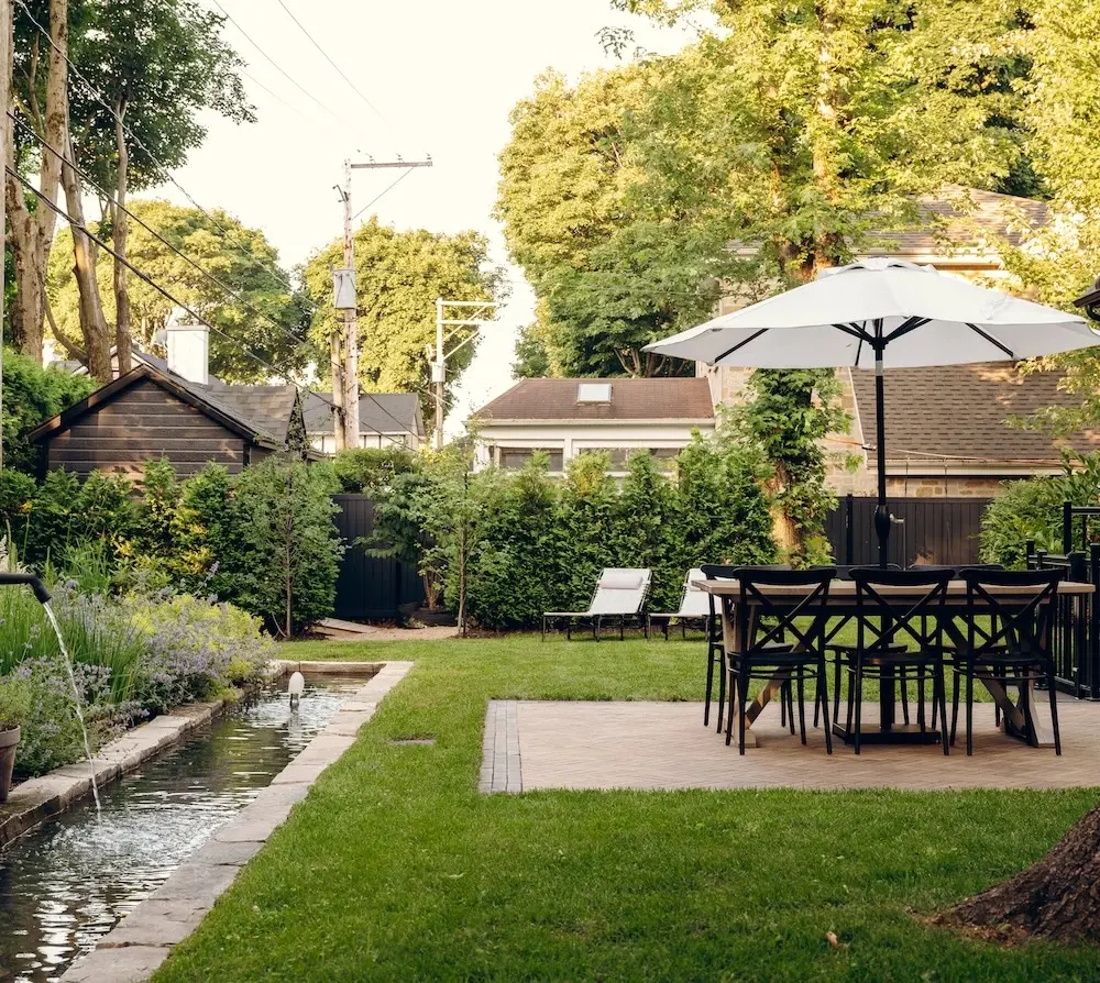After photo of a renovated backyard garden with a water feature, stepping-stone path, lawn, lounge area, dining table, umbrella, and lush greenery.