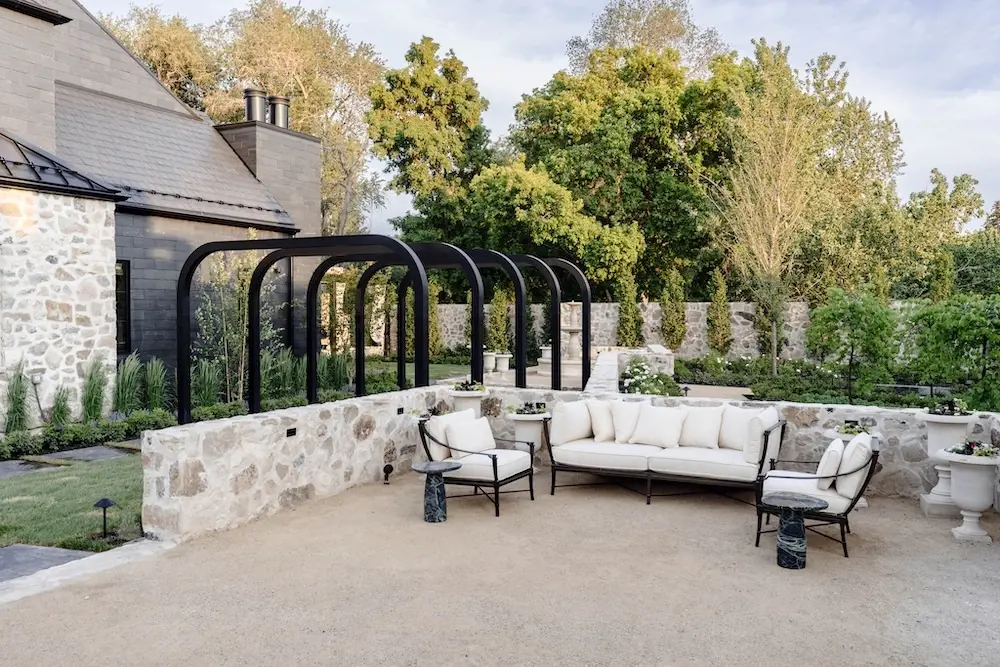 Outdoor lounge terrace with black metal arches, stone wall, cream seating, gravel-style surface, and layered garden planting.