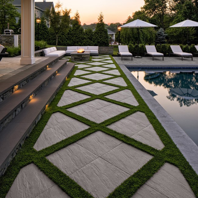 Everest Slabs by Techo-Bloc in beige, textured diamond patio with grass joints beside pool and white loungers.