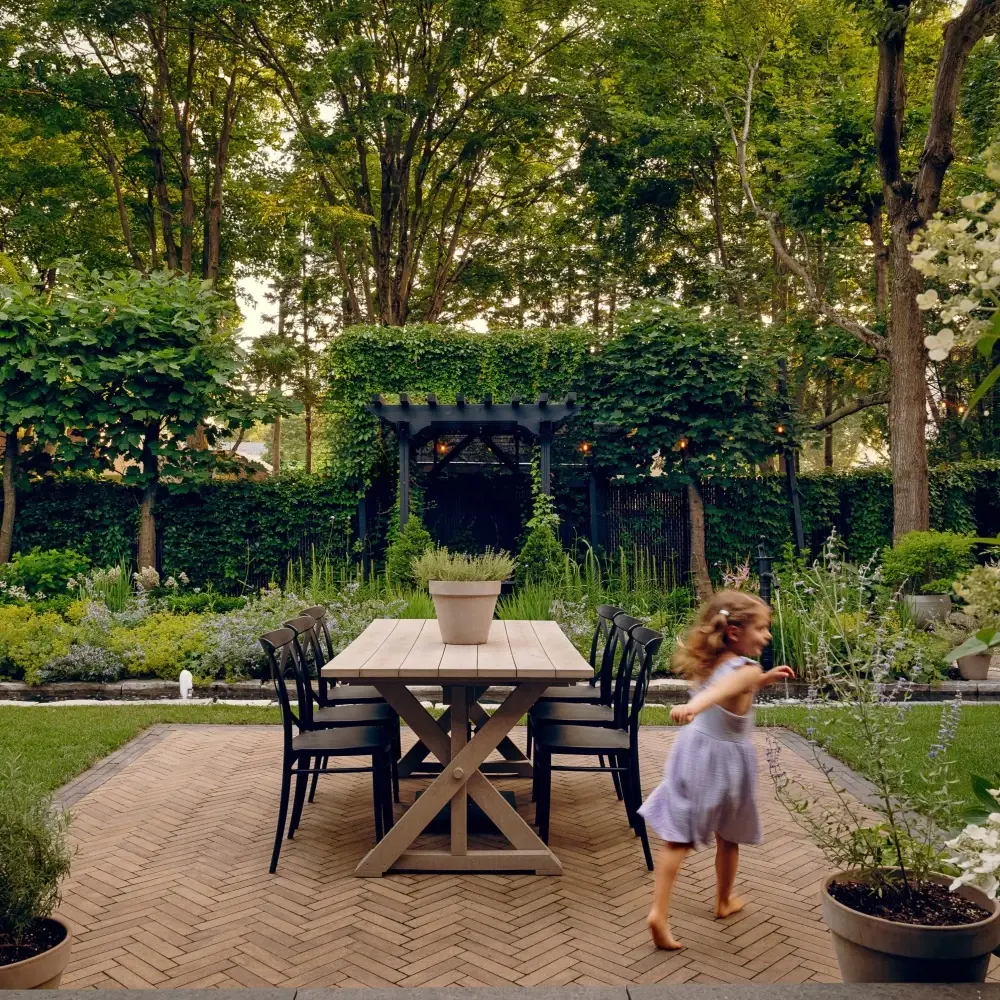 Salle à manger extérieure avec table en bois sur un patio en pavés à motif chevrons entouré d’un aménagement paysager luxuriant