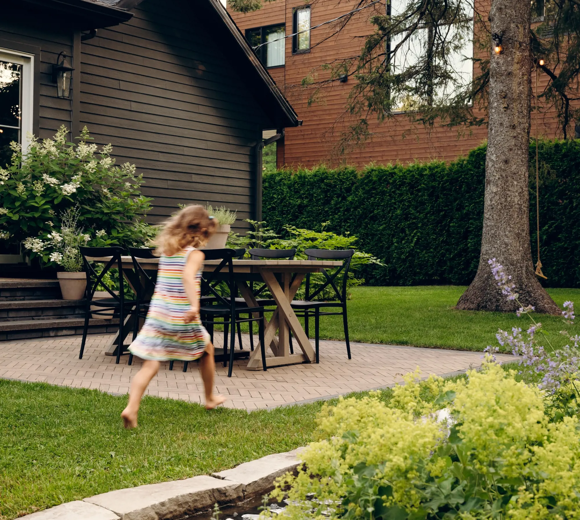 Family-friendly backyard dining area with Techo-Bloc Westmount pavers, Limestone cap edging, lawn, mature tree, hedges, and a child running beside the patio.
