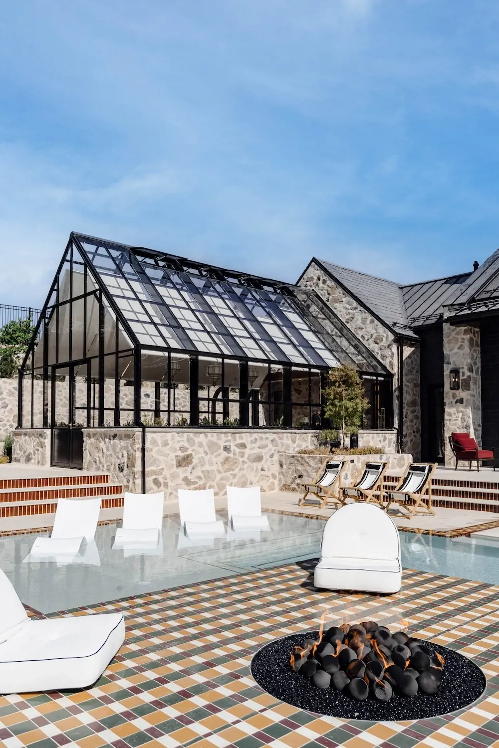 Luxury backyard pool area with a greenhouse-style structure, lounge chairs, fire feature, patterned paving, and stone architecture.