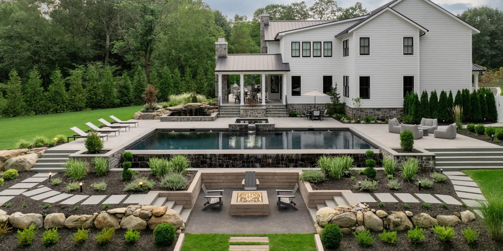 Cour arrière haut de gamme avec piscine, patio à niveaux multiples, murs de soutènement et foyer en pavés Techo-Bloc
